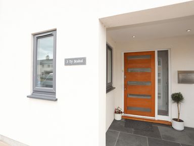 An entrance area with a wooden front door a gray window two potted plants a mailbox and a nameplate saying 3 Ty Stabal in Pwllheli
