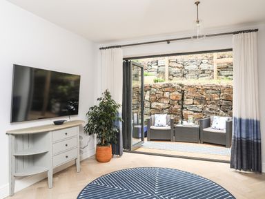 A living area with a wall-mounted television a sideboard plant and bi-fold doors leading to an outdoor seating area with chairs a table and a stone wall at 3 Ty Stabal in Pwllheli