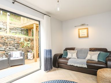 A living room with a dark leather sofa and cushions next to a sliding glass door opening to a patio with outdoor chairs at 3 Ty Stabal in Pwllheli