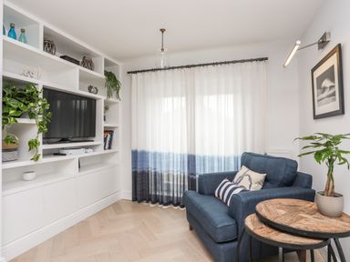 A living room with a blue armchair two wooden nested tables a built in white shelving unit with a television and plants at 3 Ty Stabal in Pwllheli