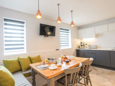 A kitchen with a wooden dining table set with breakfast items green cushions on bench seating grey chairs and appliances on counter at 3 Ty Stabal in Pwllheli