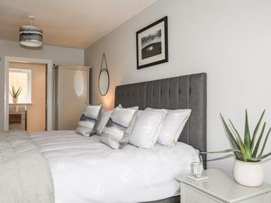 A bedroom with a grey headboard bed white bedding with cushions a wardrobe a round mirror and a plant on a bedside table at 3 Ty Stabal in Pwllheli
