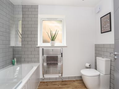A bathroom with a bathtub, a towel rack with a towel, a toilet, a window with a plant, and a framed picture at 3 Ty Stabal in Pwllheli