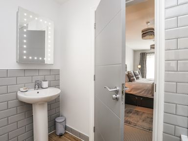 A bathroom with a sink mirror and trash bin next to a partially open door showing a bedroom with a bed, pillows, lamp, and window at 3 Ty Stabal in Pwllheli