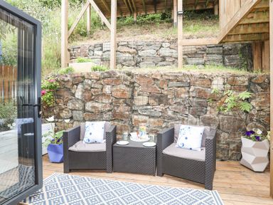 An outdoor seating area with two armchairs and a table on a wooden deck with potted plants and a stone retaining wall at 3 Ty Stabal Pwllheli