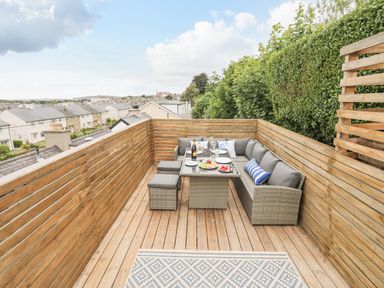 An outdoor wooden deck with a wicker dining set including a sofa table and stools overlooking a neighborhood at 3 Ty Stabal in Pwllheli