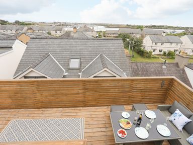 A rooftop terrace with wooden flooring and railing a table set with plates glasses and food with rooftops and a marina in the background at 3 Ty Stabal in Pwllheli