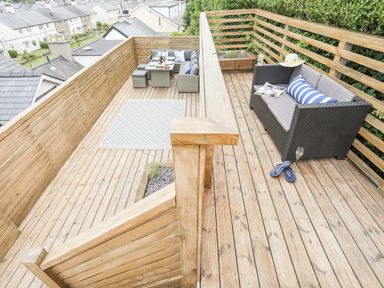 A wooden outdoor deck with seating areas including a sofa with striped pillow and a dining table with chairs at 3 Ty Stabal in Pwllheli