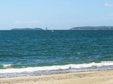 A beach with waves and two sailboats on the sea with islands in the distance at 3 Ty Stabal in Pwllheli