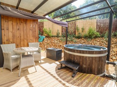 An outdoor covered patio with two wicker chairs a round table a wooden hot tub and a garden hose reel at The Shepherds Hut at Bridge Lake Farm & Fishery in Chacombe