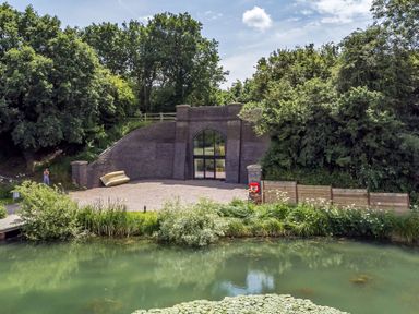A brick structure with arched glass doors surrounded by trees and a pond in front at The Shepherds Hut at Bridge Lake Farm & Fishery in Chacombe