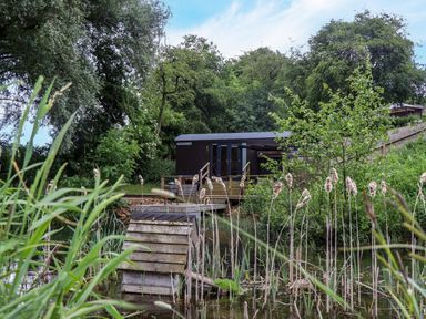 A wooden cabin near a pond surrounded by trees and plants at The Shepherds Hut at Bridge Lake Farm & Fishery in Chacombe