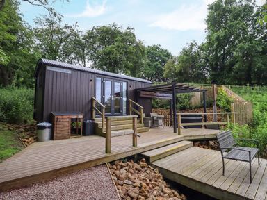 An outdoor wooden deck with a bench and seating area next to a small dark brown hut surrounded by trees at The Shepherds Hut at Bridge Lake Farm & Fishery in Chacombe