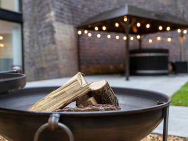 A fire pit with logs in the foreground and a gazebo with string lights in a backyard at The Shepherds Hut at Bridge Lake Farm & Fishery Chacombe