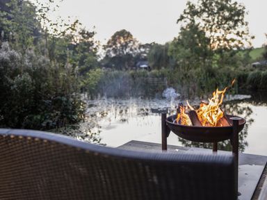 A fire pit with burning wood beside a pond with plants and trees in the background at The Shepherds Hut at Bridge Lake Farm & Fishery in Chacombe