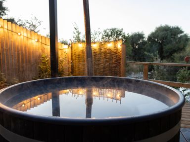 A round outdoor hot tub with steam above the water on a wooden deck with string lights on a fence at The Shepherds Hut at Bridge Lake Farm & Fishery in Chacombe