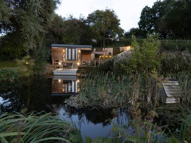 A wooden hut with stairs and outdoor seating near a pond surrounded by trees and plants at The Shepherds Hut at Bridge Lake Farm & Fishery in Chacombe