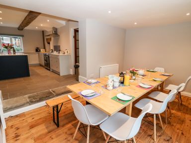 A dining area with a wooden table set for breakfast and white chairs next to a kitchen with light cabinets at Ley Mill Farm in Huntley