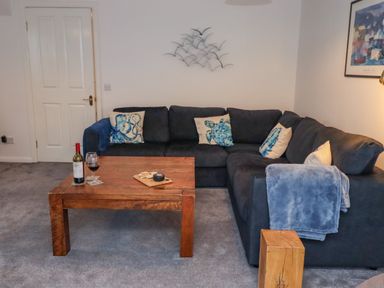 A living room with a dark sectional sofa with blue and white cushions a wooden coffee table with wine and cheese and a wooden side table at Pond Farm View in Hinderwell
