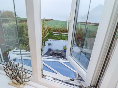 View through an open window overlooking a patio with a round wooden table bench set and potted plants surrounded by greenery at Pond Farm View in Hinderwell