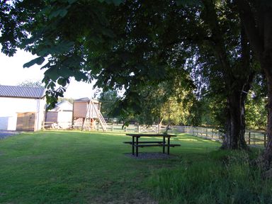 A yard with a picnic table under trees and a wooden play structure with a fenced area at Dinefwr Cottage in Golden Grove near Llandeilo