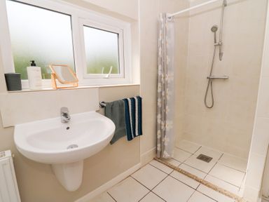 A bathroom with a wall-mounted sink a towel rack with two towels a shower area with a floral shower curtain and a frosted window at Dinefwr Cottage in Golden Grove near Llandeilo