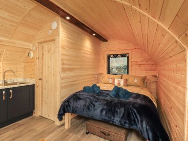 A wooden cabin interior with a bed covered in animal print bedding and towels next to a small kitchenette at The Stag - Crossgate Luxury Glamping in Hartsop near Glenridding
