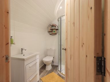 A bathroom with a toilet, vanity with sink, round wall shelf with decor, curved glass shower, and wooden door at The Stag - Crossgate Luxury Glamping in Hartsop near Glenridding