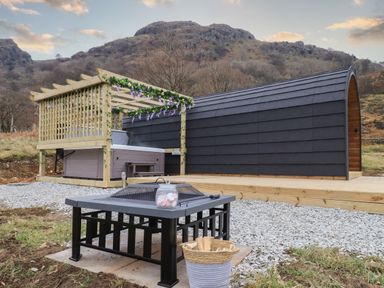 A black pod-shaped cabin with a wooden deck a hot tub under a pergola decorated with flowers a fire pit with marshmallows on it and a basket of firewood outdoors at The Stag - Crossgate Luxury Glamping in Hartsop near Glenridding