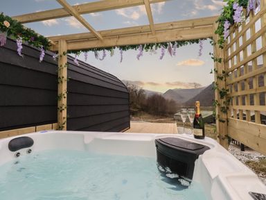 An outdoor hot tub with champagne bottle and glasses under a wooden pergola with flowers at The Stag - Crossgate Luxury Glamping in Hartsop near Glenridding