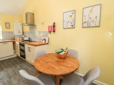 A kitchen with a wooden round table and four gray chairs a bowl with snacks on the table cabinets with wood countertops an oven toaster and wall art at Kizzie Cottage Guyzance near Warkworth