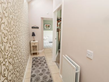 A narrow hallway with patterned wallpaper on one side a small table with a lamp a rug on the floor and an open doorway showing a room with chairs at Kizzie Cottage Guyzance near Warkworth