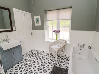 A bathroom with a bathtub a sink with a mirror a white wicker chair and a window with a vase of flowers at Kizzie Cottage in Guyzance near Warkworth