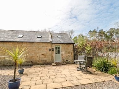 A stone cottage with a paved patio featuring two potted plants and a table with two chairs at Kizzie Cottage in Guyzance near Warkworth