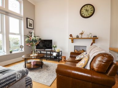 A living room with a leather sofa, a TV on a stand, a round coffee table with flowers, and a wall clock at King Edward's View in Newquay