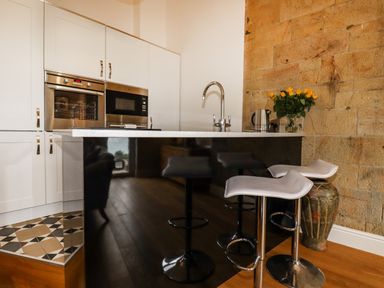 A kitchen with white cabinets an oven and microwave a black island with a sink and two white bar stools a kettle and flowers in a vase by a stone wall at King Edward's View in Newquay