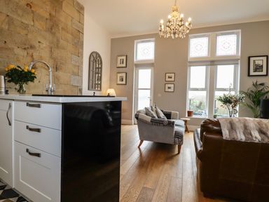 A living room with sofas, a chandelier, and a kitchen island with a sink and flowers at King Edward's View in Newquay