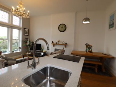 A kitchen island with a sink and stovetop overlooking a living area with a sofa table and TV at King Edward's View in Newquay