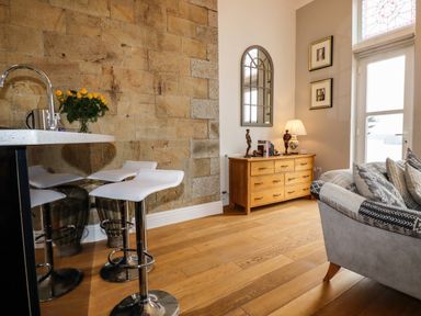 A kitchen counter with stools and a vase of yellow flowers beside a wooden chest of drawers and a sofa at King Edward's View in Newquay