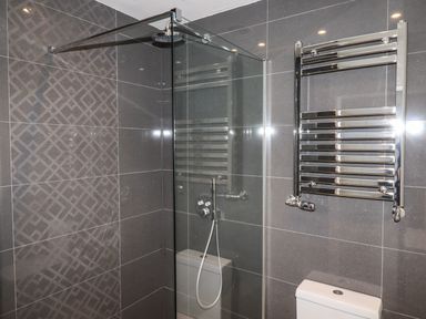 A bathroom with gray tiled walls a glass shower screen a rainfall showerhead a white toilet and a chrome towel radiator at King Edward's View in Newquay
