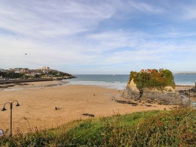 A sandy beach with a rock formation with a house on top near the sea and buildings in the distance at King Edward's View in Newquay