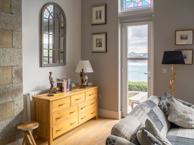 A living room corner with a wooden dresser with books and statues next to a window overlooking water at King Edward's View in Newquay