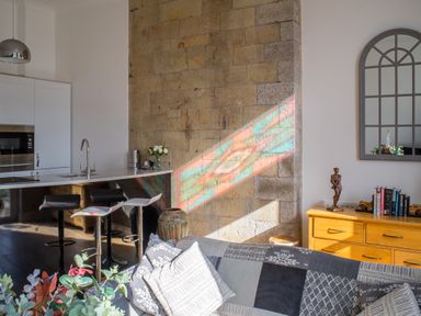 A kitchen with a white counter and bar stools stone wall with colorful light reflection and a wooden dresser with books and a small statue at King Edward's View in Newquay