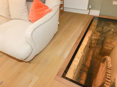 A corner of a beige sofa with an orange cushion on a wooden floor next to a glass floor panel revealing old stone and a water wheel at The Old Watermill in St Mabyn