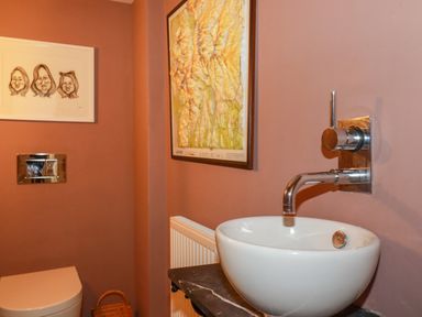 A bathroom with a white sink mounted on a dark counter a wall-mounted faucet a toilet a wicker basket and framed pictures on the wall at The Old Watermill in St Mabyn