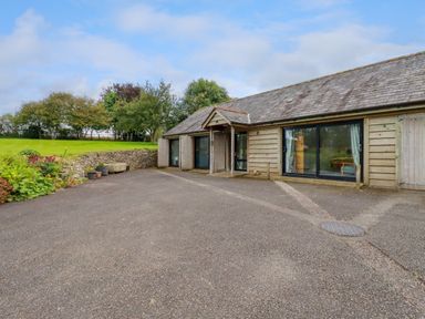 A wooden building with large glass sliding doors and a small entrance canopy next to a paved area with greenery and stone wall at The Old Watermill in St Mabyn