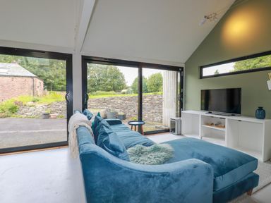 A living room with a blue sofa and a TV on a white stand with large glass doors showing an outdoor stone wall at The Old Watermill in St Mabyn