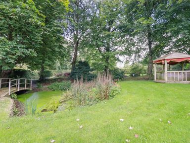 A garden with a small wooden bridge over a pond and a wooden gazebo surrounded by trees at The Old Watermill in St Mabyn
