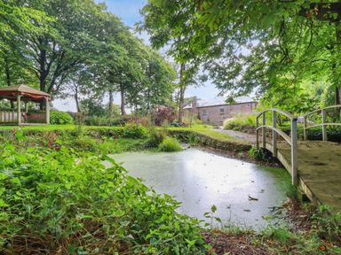 A garden with a small pond a wooden bridge trees and a gazebo near a brick house at The Old Watermill in St Mabyn