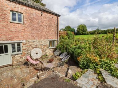 A stone patio with outdoor chairs and a fire pit next to a brick and stone house with greenery at The Old Watermill in St Mabyn